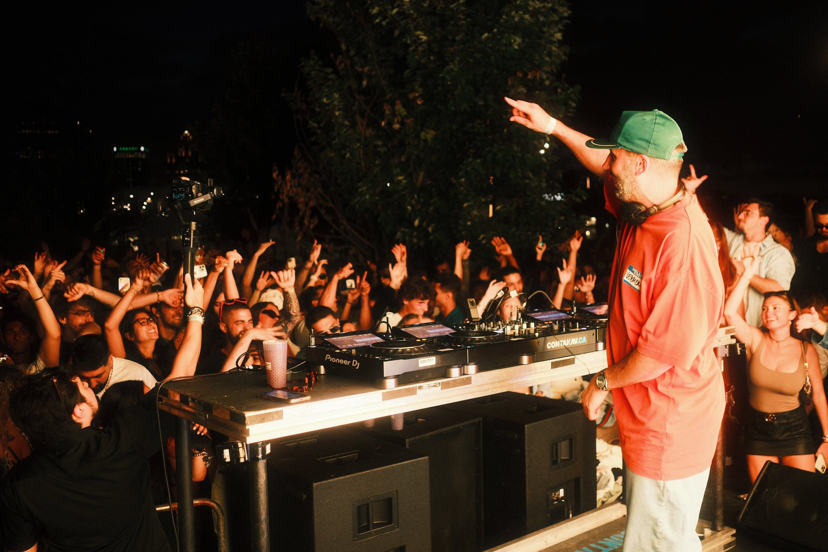 DJ in a green cap pointing to an outdoor crowd at dusk